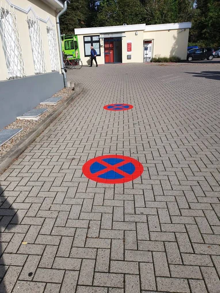 Gepflasterte Fläche mit drei aufgemalten roten und blauen „Parken verboten“-Symbolen in der Nähe eines Gebäudes.