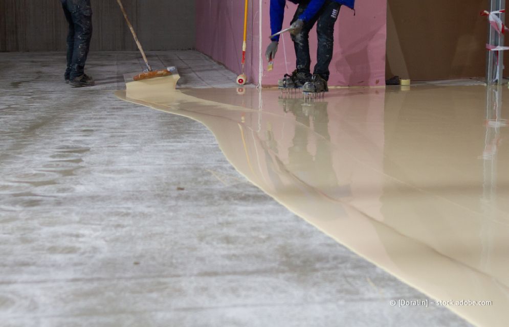 Arbeiter tragen mit Besen und Walzen eine helle Beschichtung auf einen Betonboden auf.