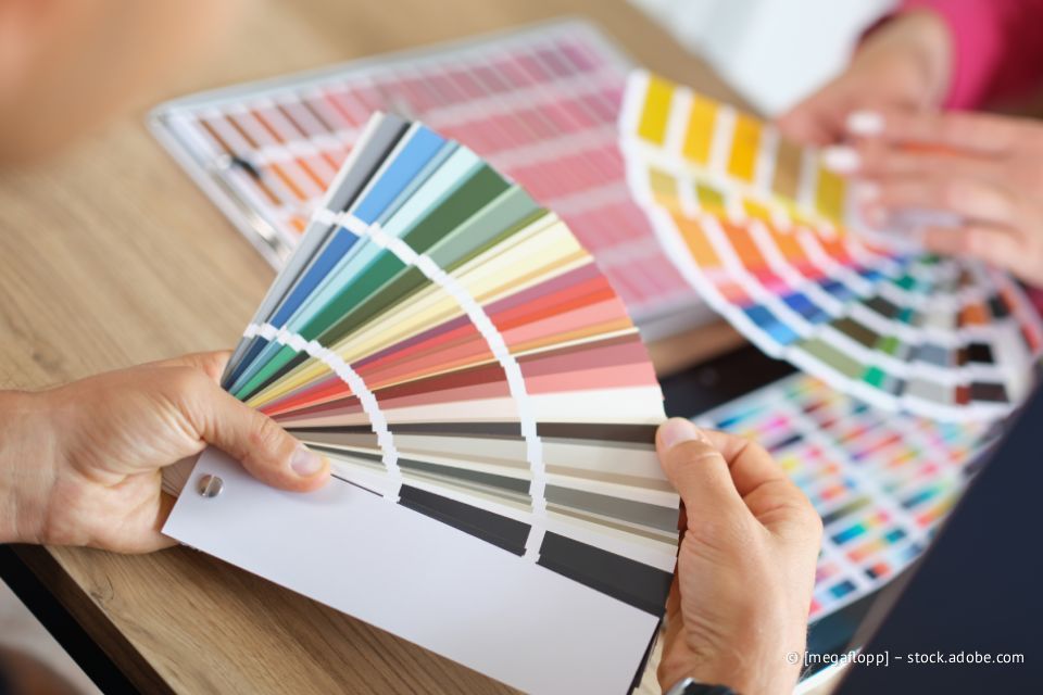 Hände halten Farbmuster und wählen Farben aus. Weitere Farbmuster liegen auf einem Tisch.