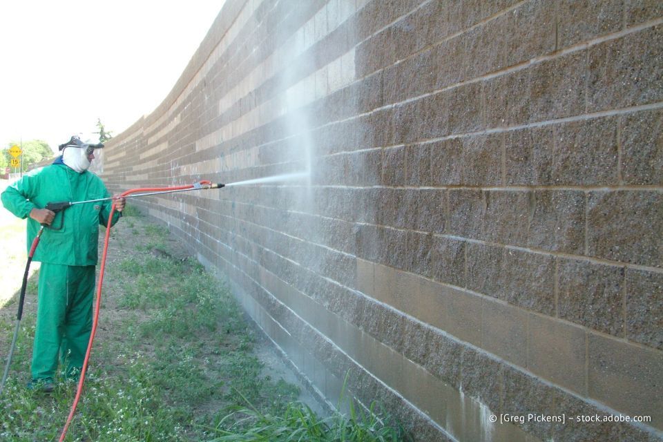 Eine Person in grüner Schutzkleidung reinigt im Freien mit einem Hochdruckreiniger eine lange, hellbraune Backsteinmauer.