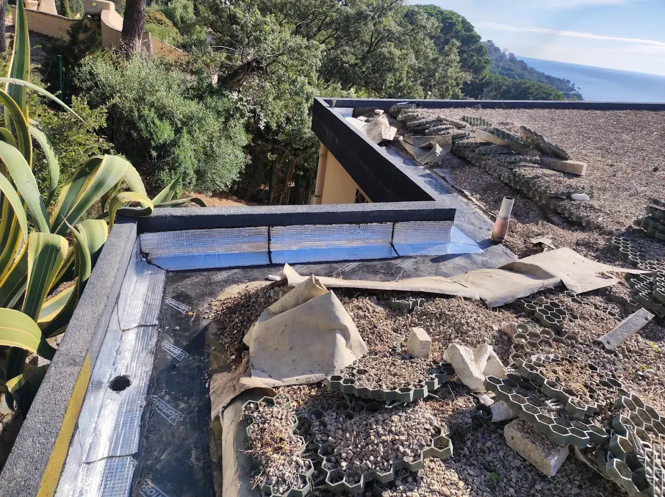 Un toit plat recouvert de gravier abîmé et bordé de solins étanches argentés, avec vue sur les arbres et la mer.