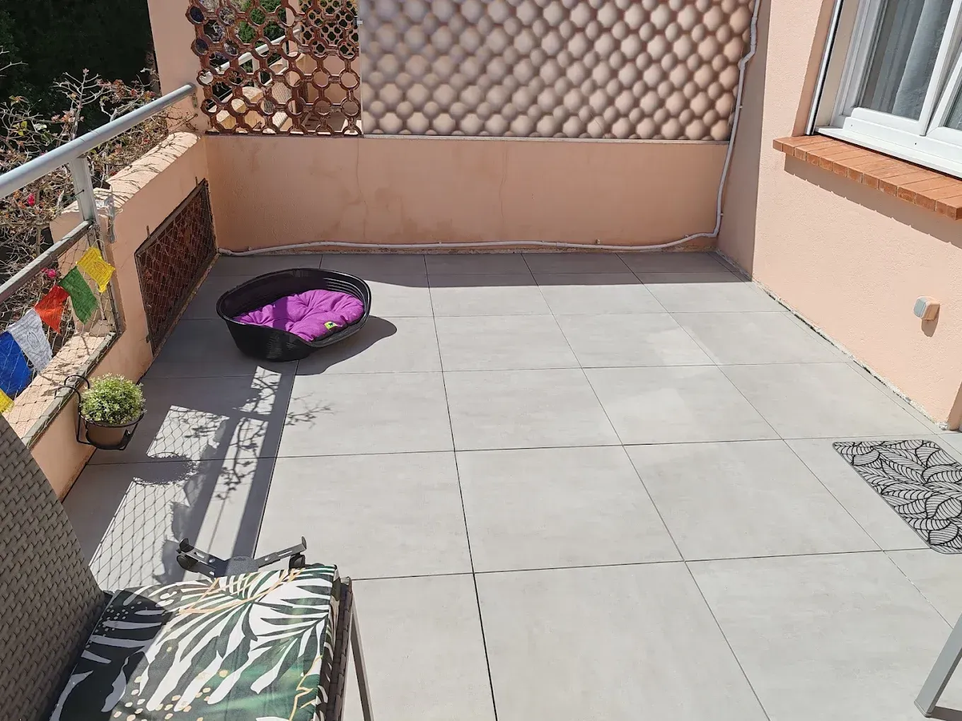 Un balcon ensoleillé avec un carrelage gris au sol, un panier pour animaux avec un coussin violet, une plante en pot et un fauteuil d'angle avec des coussins.