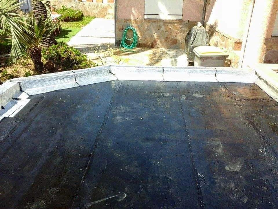 Une surface de toit plate, recouverte de bitume noir et bordée de solins métalliques blancs, avec un jardin résidentiel en arrière-plan.