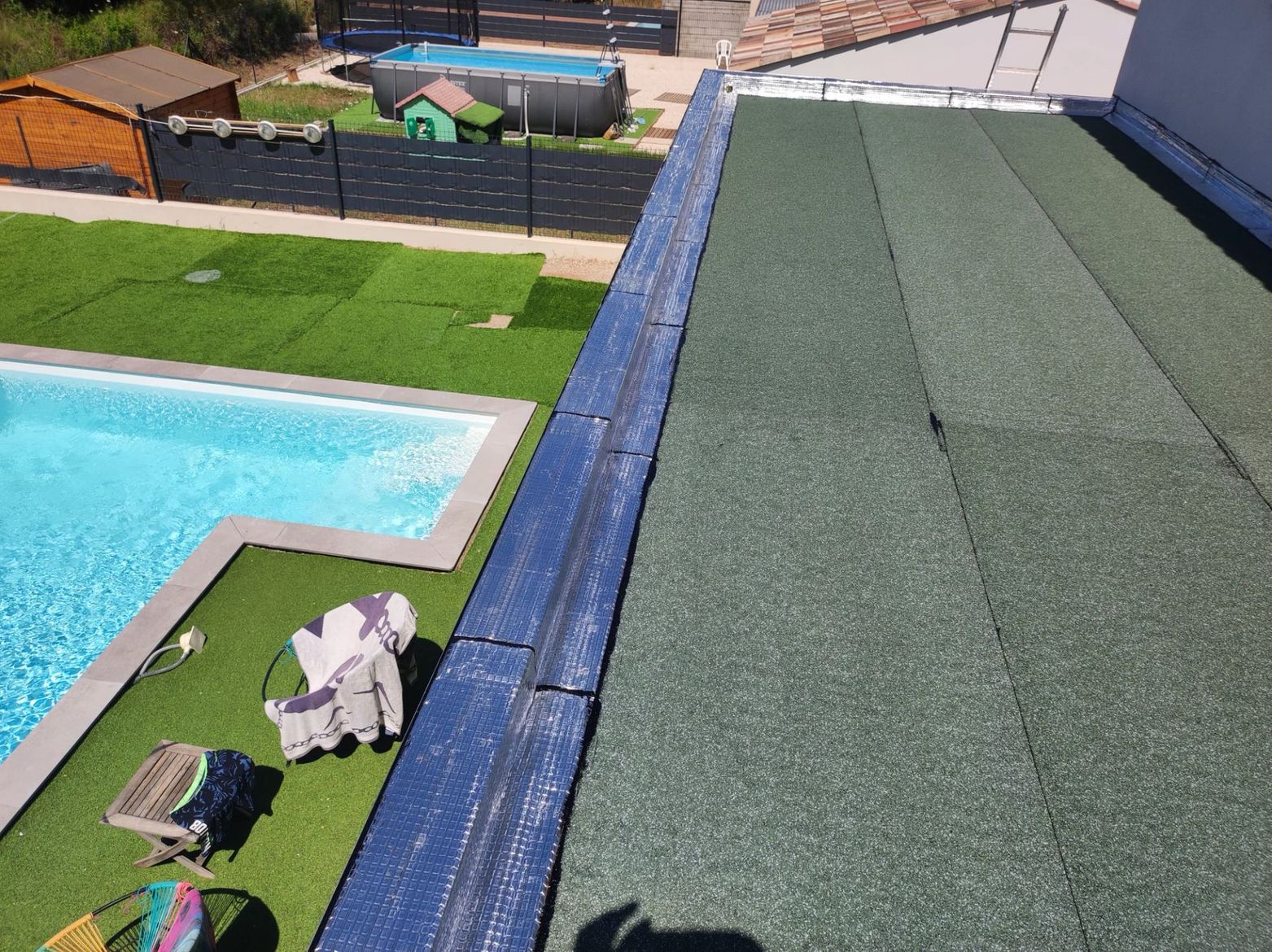 Vue en plongée d'un toit plat recouvert de feutre minéral vert foncé, donnant sur un jardin avec piscine et pelouse.