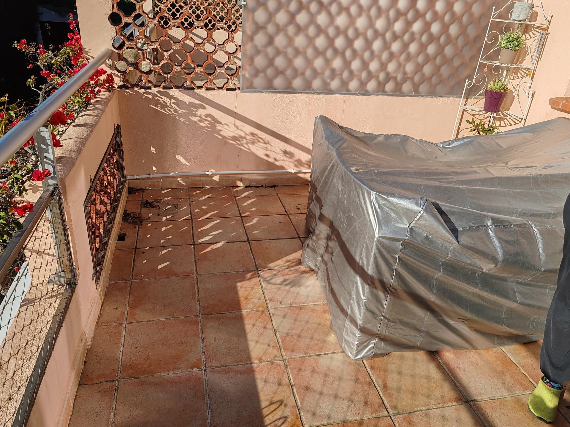 Un balcon carrelé est aménagé avec un grand meuble recouvert d'une bâche argentée, des plantes en pot et une rambarde en métal.