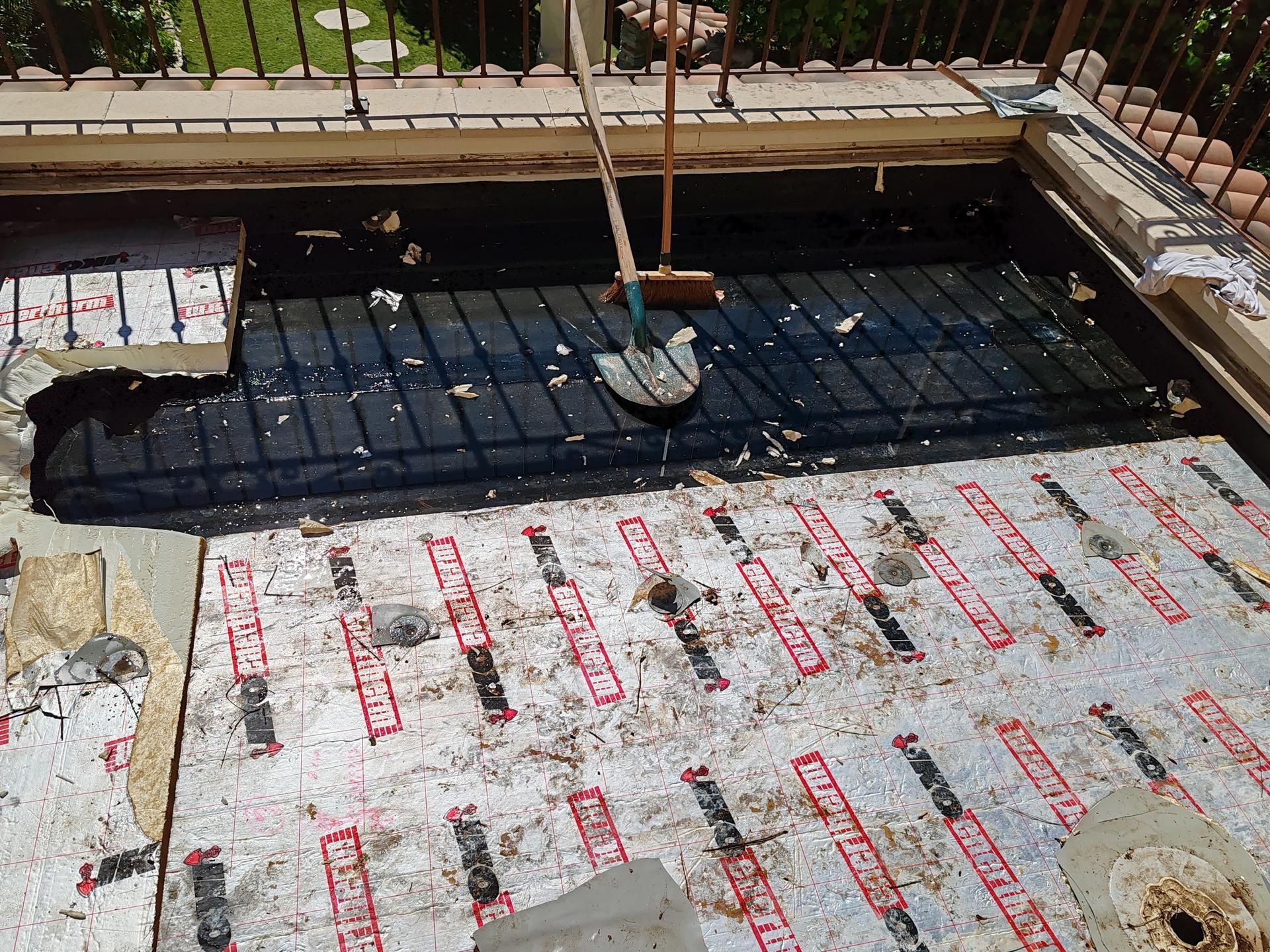 Zone de construction avec une membrane d'étanchéité noire sous des panneaux isolants sur une terrasse de balcon, avec une pelle au milieu.