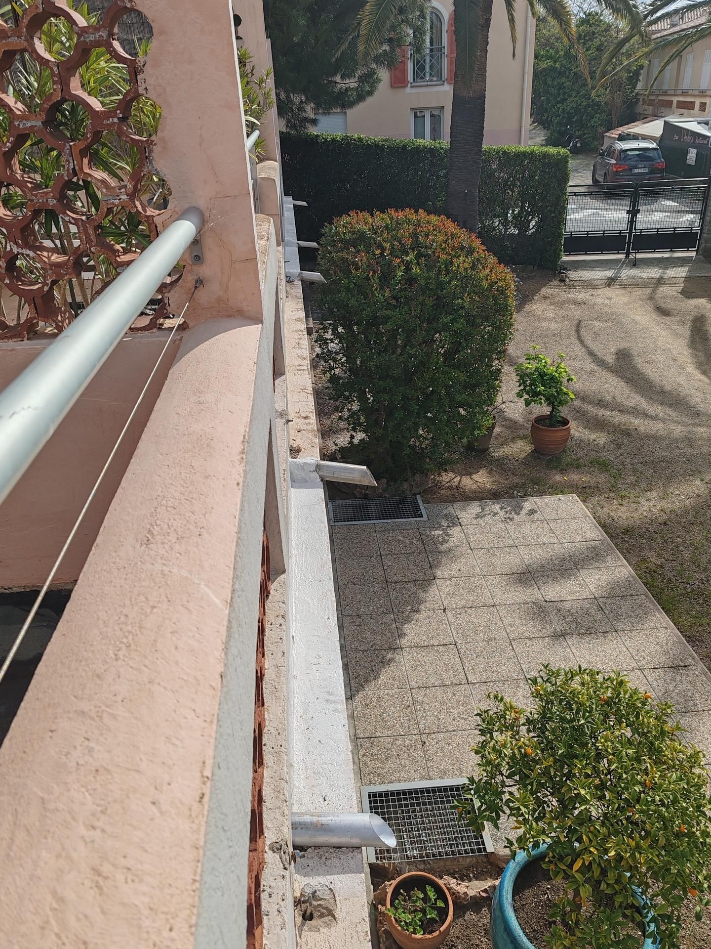 Vue en plongée d'un rebord de balcon donnant sur une cour gravillonnée avec des buissons, une plante en pot et une voiture garée.