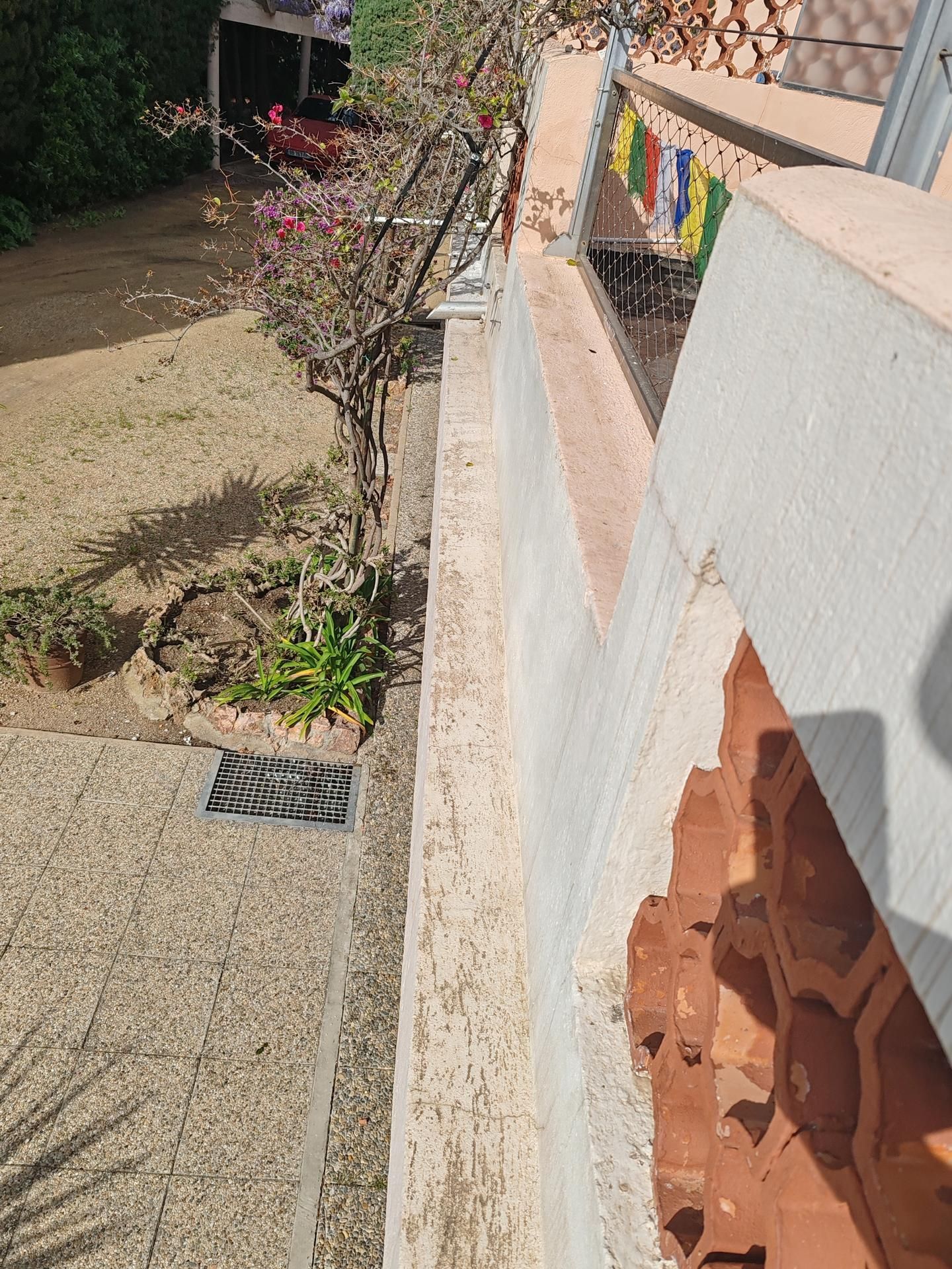 Une étroite corniche en béton jouxte une terrasse carrelée, avec une plante et une petite grille d'égout au sol à proximité.