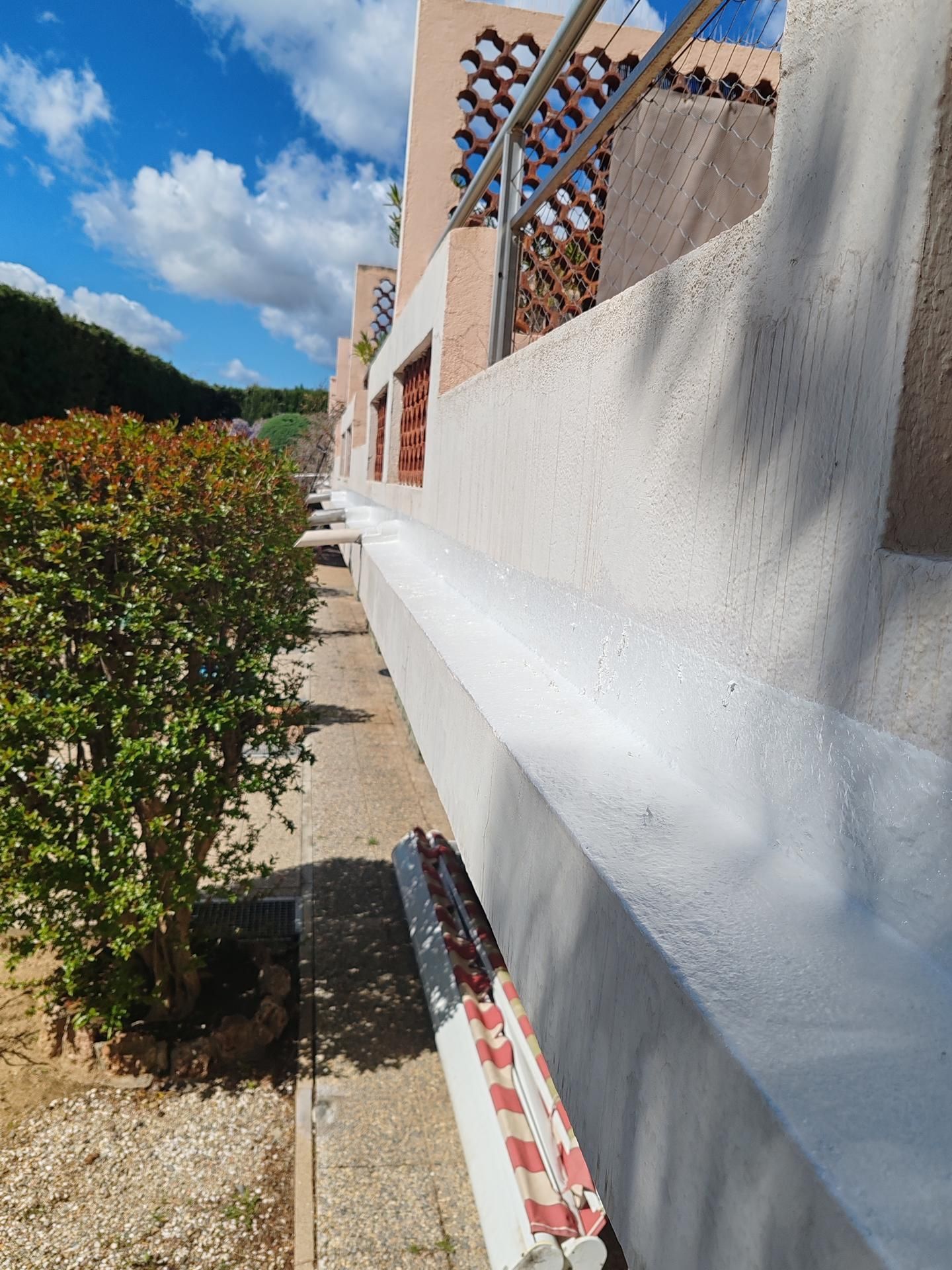 Un mur blanc texturé longe un chemin de gravier, à côté d'un arbuste vert.