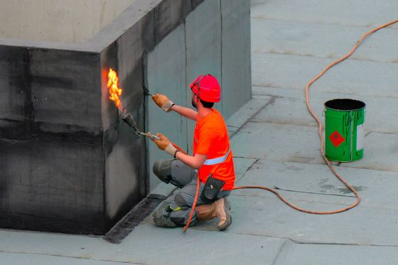 Un ouvrier vêtu d'une chemise orange et d'un casque rouge utilise un chalumeau pour sceller le matériau de toiture d'un bâtiment.