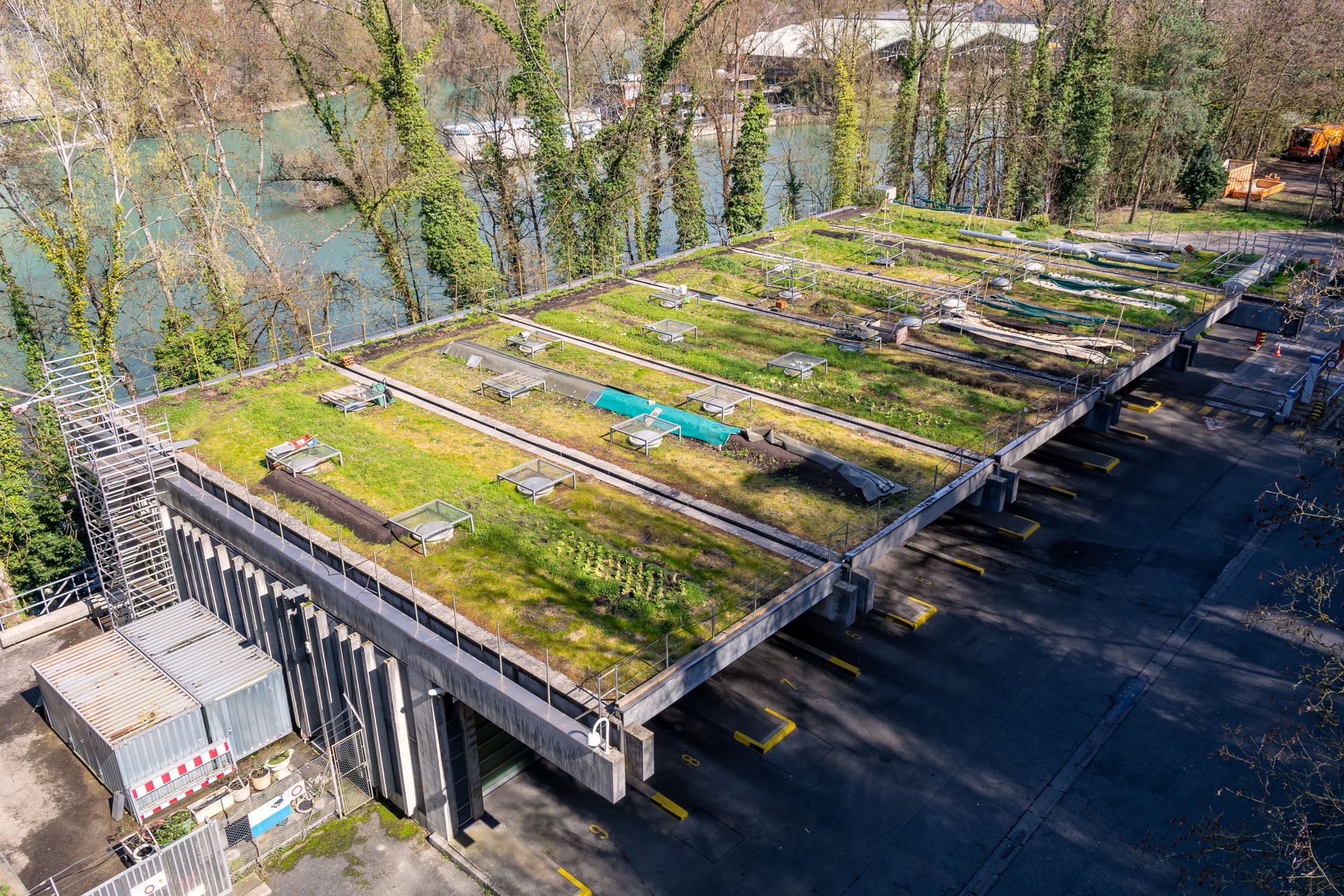Vue aérienne d'une longue structure en béton avec un toit végétalisé, située au bord d'une rivière et d'une zone boisée.