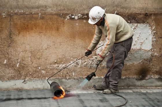Un ouvrier du bâtiment utilise un chalumeau à propane pour chauffer et appliquer une couche de matériau imperméabilisant sur une surface en béton.