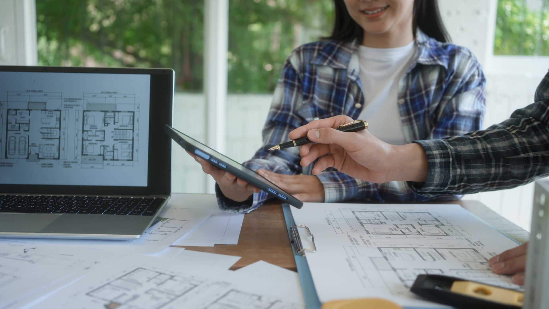 Deux personnes examinent des plans architecturaux en pointant du doigt une tablette posée sur un bureau.