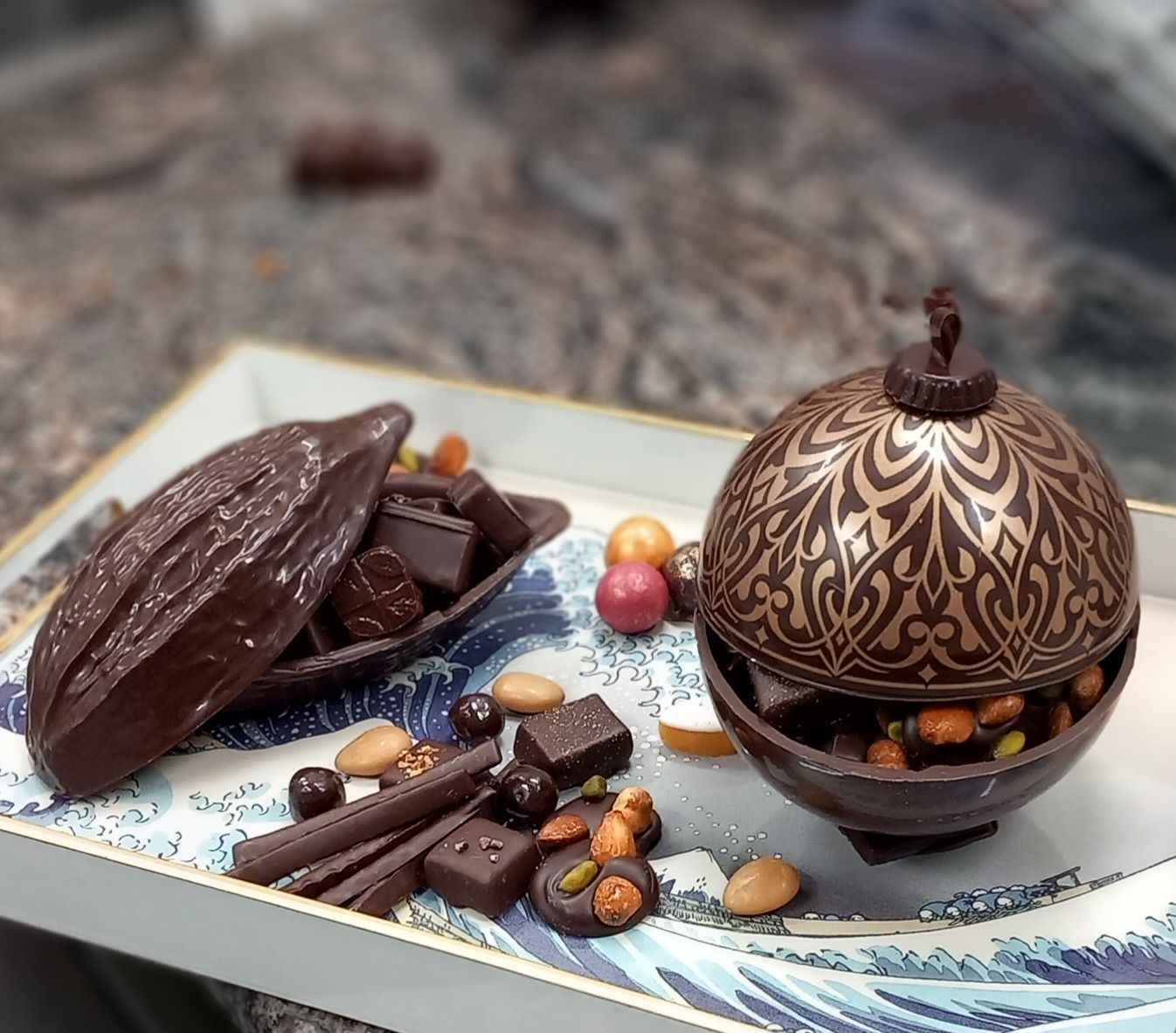 Des friandises au chocolat disposées avec art sur un plateau : cabosse de cacao, sphère ornée, noix, bonbons, marron foncé et or.