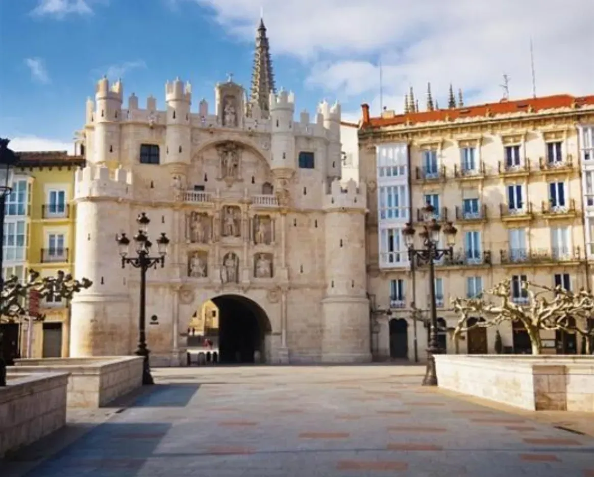 Entrada con arco de piedra con tallas ornamentales, flanqueada por edificios en una plaza soleada.