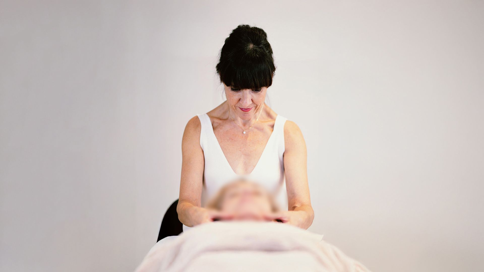 Una mujer le da un masaje en la cabeza a otra mujer. Ambas están en una habitación blanca.