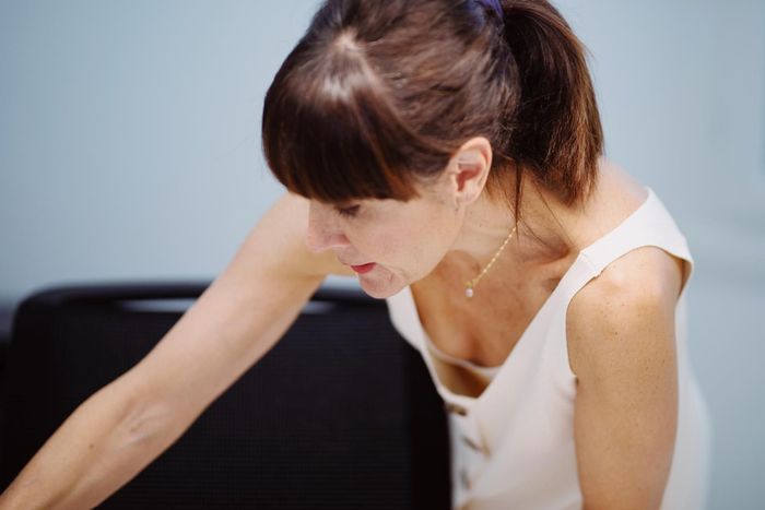 Una mujer, vestida con un vestido de color claro, se inclina hacia adelante y mira hacia abajo con expresión concentrada.