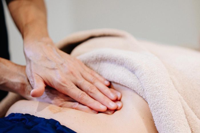 Manos masajeando la espalda de una persona, parcialmente cubierta por una manta color crema, en un primer plano.