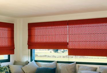 Cortinas romanas rojas estampadas instaladas en las ventanas sobre un sofá con cojines grises.