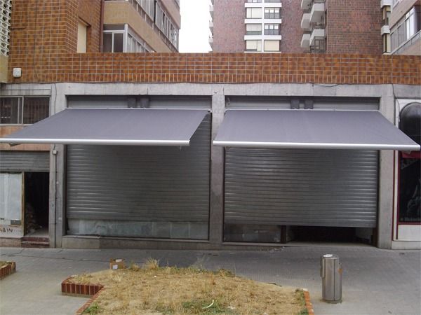 Un escaparate con dos persianas metálicas enrollables de seguridad cubiertas por toldos de tela gris a juego.