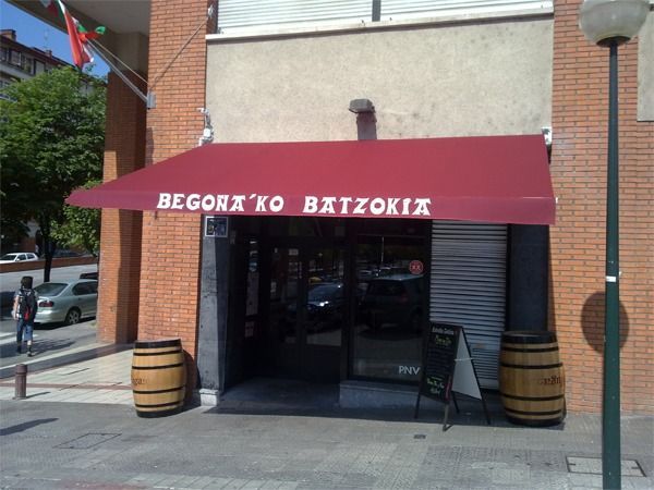 El exterior del edificio Begoña'ko Batzokia presenta un toldo rojo, una puerta oscura y dos barriles de madera en la entrada.