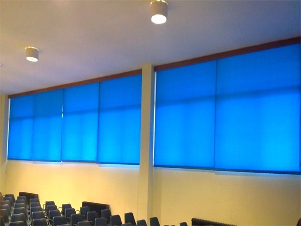 Un aula de conferencias con persianas enrollables azules que cubren una gran pared de ventanas sobre filas de sillas azules.