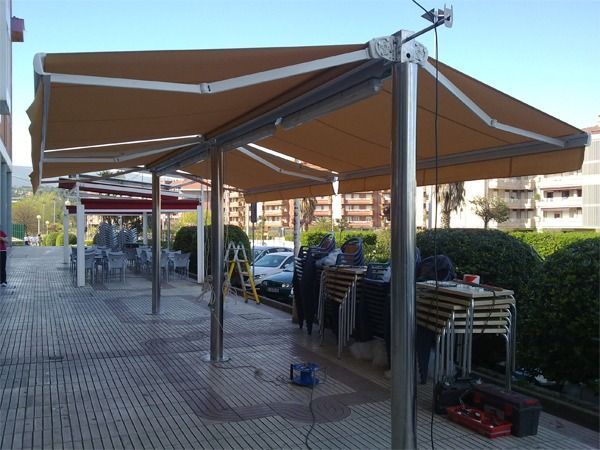 Un toldo retráctil de color beige sobre una estructura metálica que cubre un área al aire libre con mesas 
