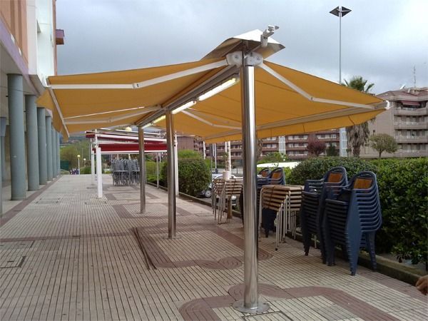 Toldos amarillos de doble cara que se extienden sobre sillas de exterior en una terraza pavimentada junto a un edificio.