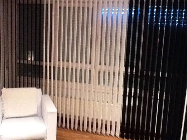 Un sillón blanco se encuentra frente a una ventana cubierta por persianas verticales con paneles blancos y negros alternados.