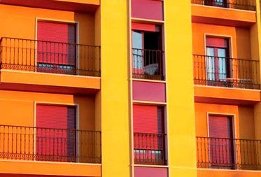 Fachada de un edificio con paneles verticales amarillos entre balcones, paredes naranjas y contraventanas rojas.