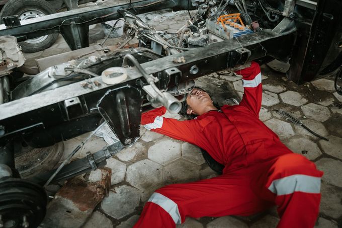 Un mecánico con un mono rojo tumbado en el suelo bajo el chasis de un vehículo para realizar reparaciones.