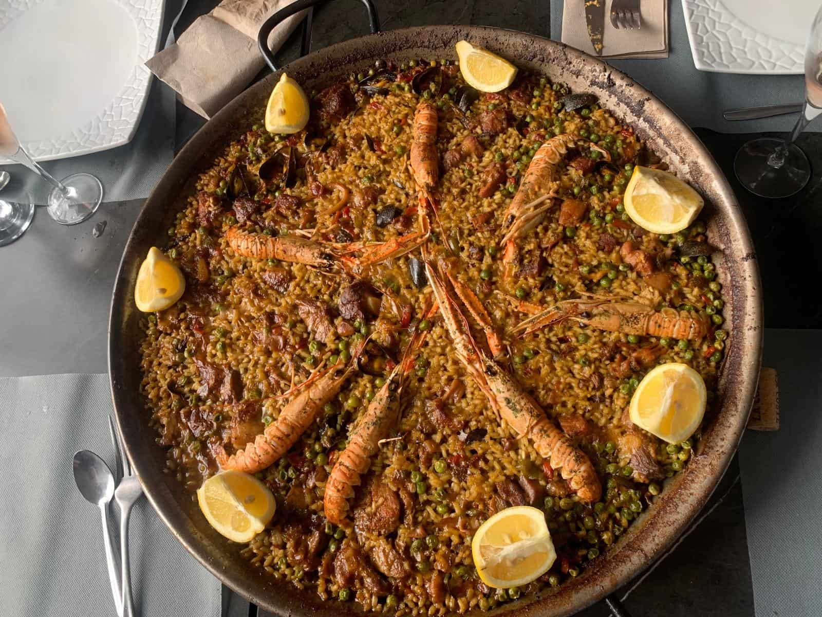 Paella en una sartén grande, adornada con camarones y rodajas de limón sobre una mesa.