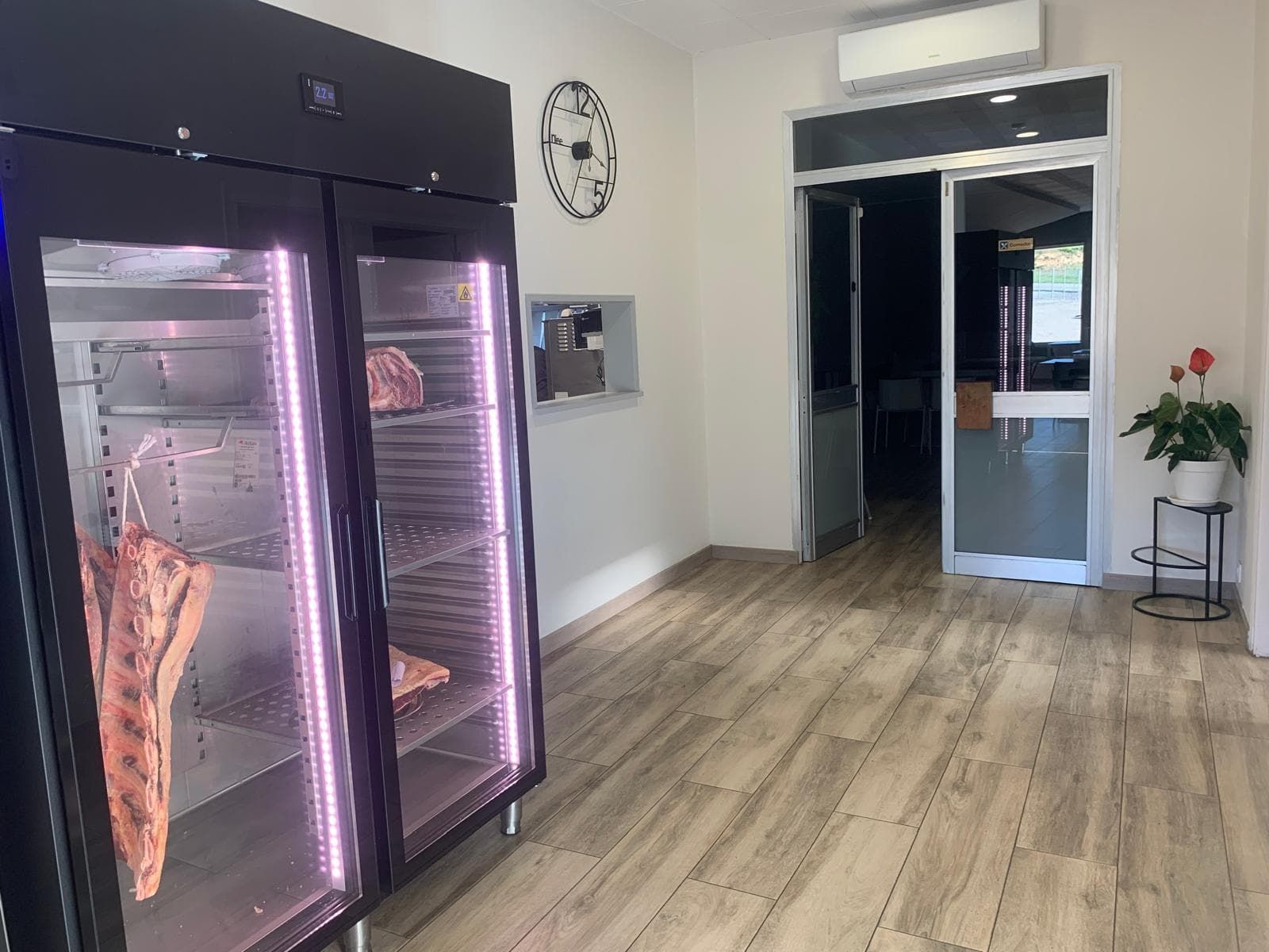 Refrigeradores negros con carne expuesta en una carnicería. Suelos y puerta de madera visibles.
