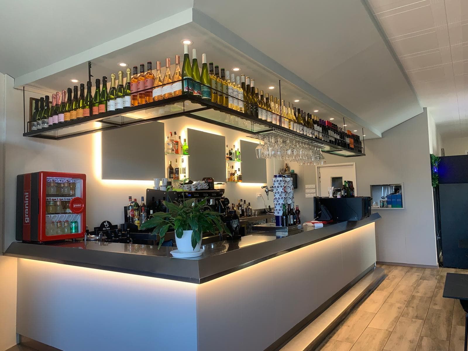 Barra con estantería superior para botellas, mostrador y pared trasera iluminados, vasos colgados.