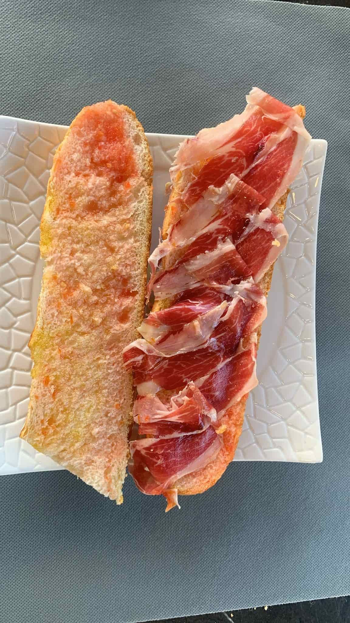 Pan cubierto con tomate y jamón curado en lonchas sobre un plato blanco.