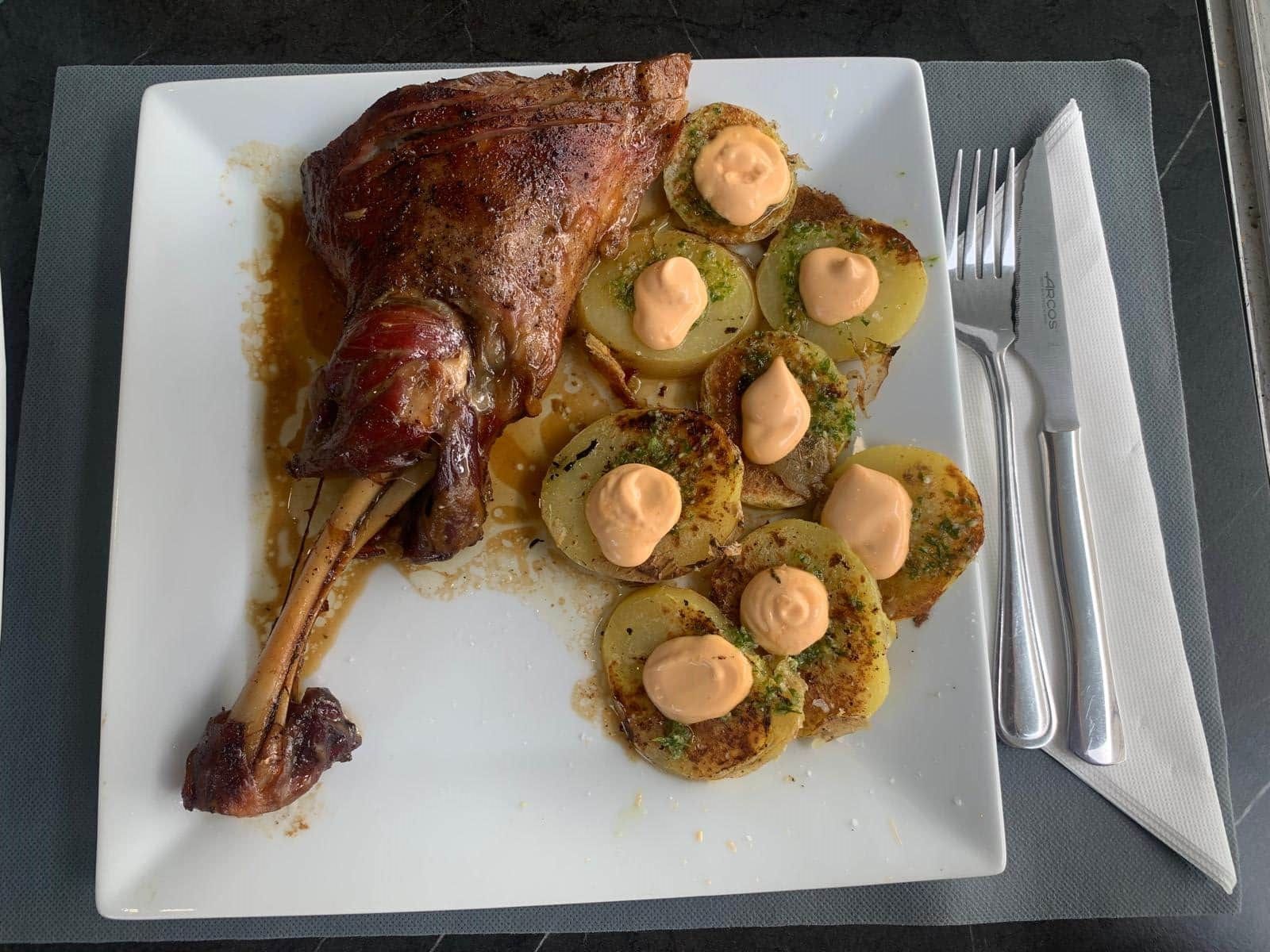 Muslo de pato asado con patatas y salsa en un plato blanco con cubiertos.