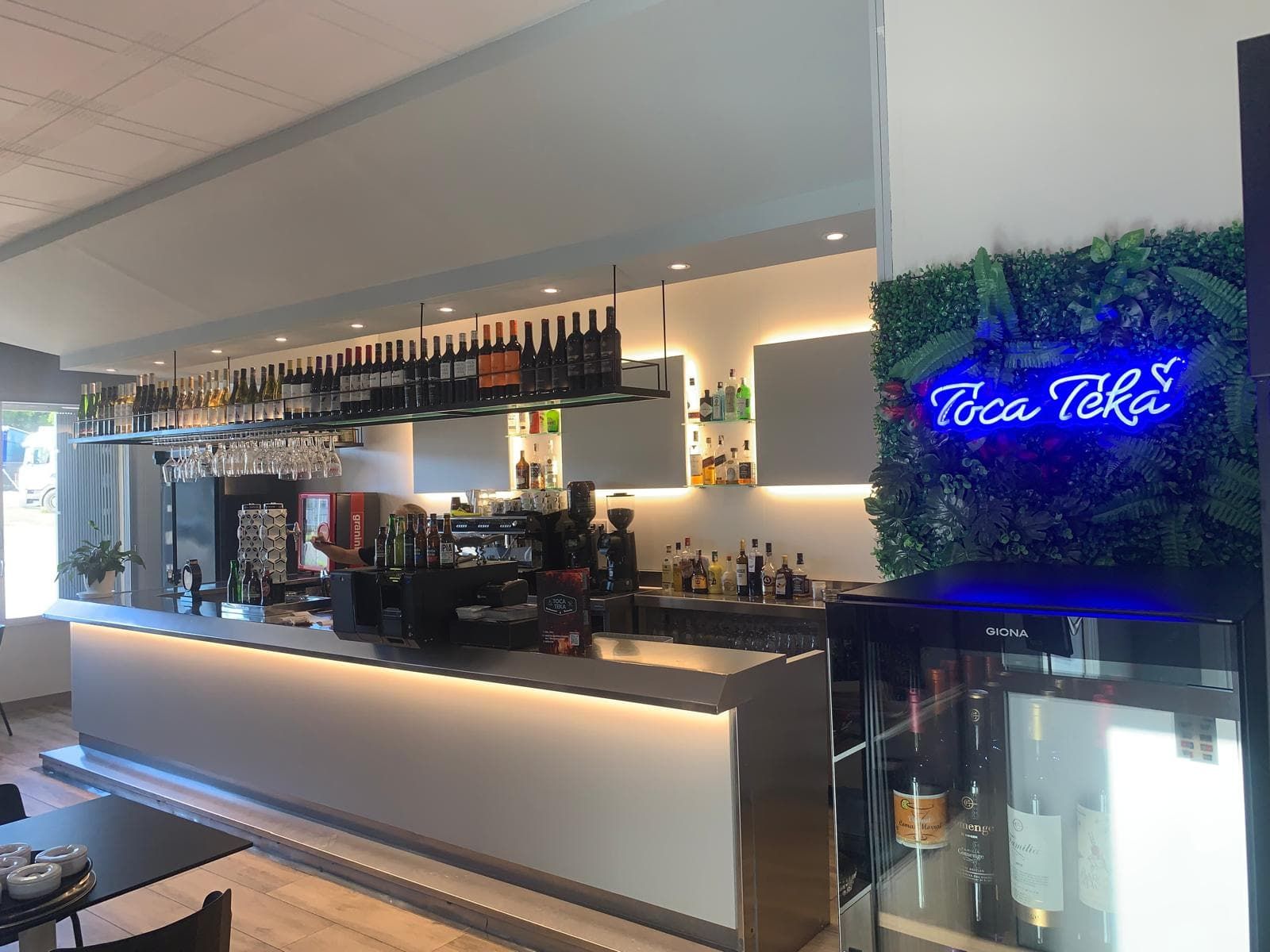 Interior de un bar, con barra iluminada, botellas y un letrero de neón que dice