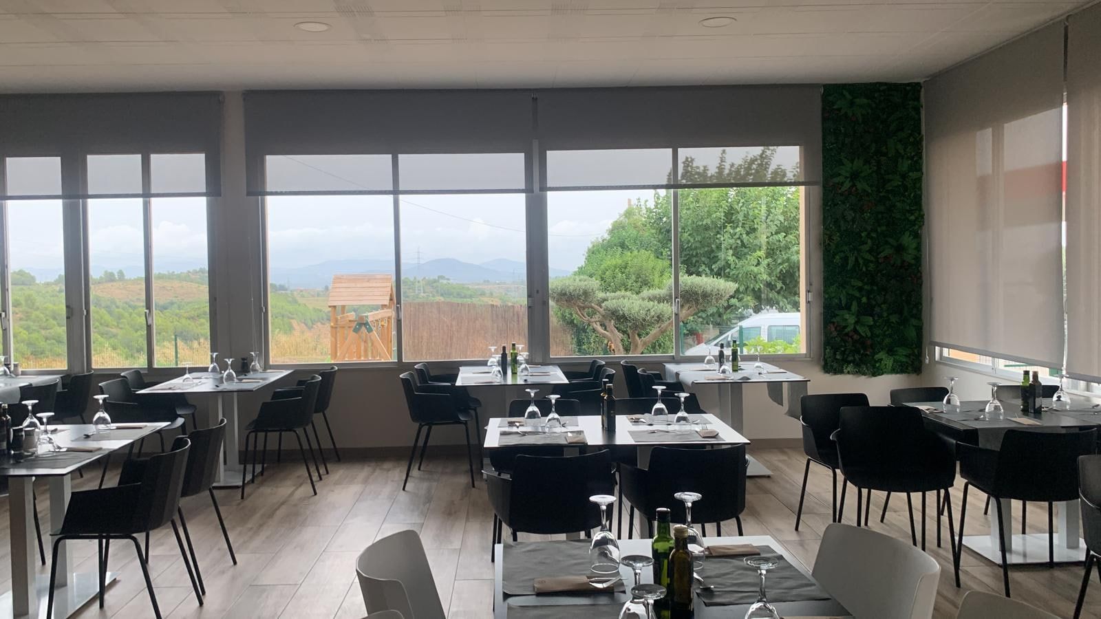 Interior de restaurante con mesas preparadas para cenar, grandes ventanales con vista panorámica y una pared verde.