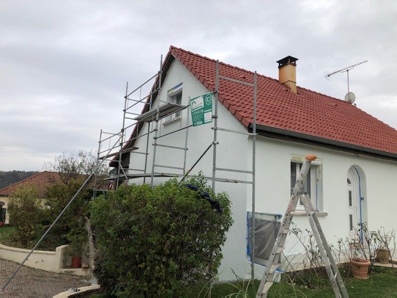 Des échafaudages sont dressés contre une maison blanche au toit rouge, sous un ciel couvert.