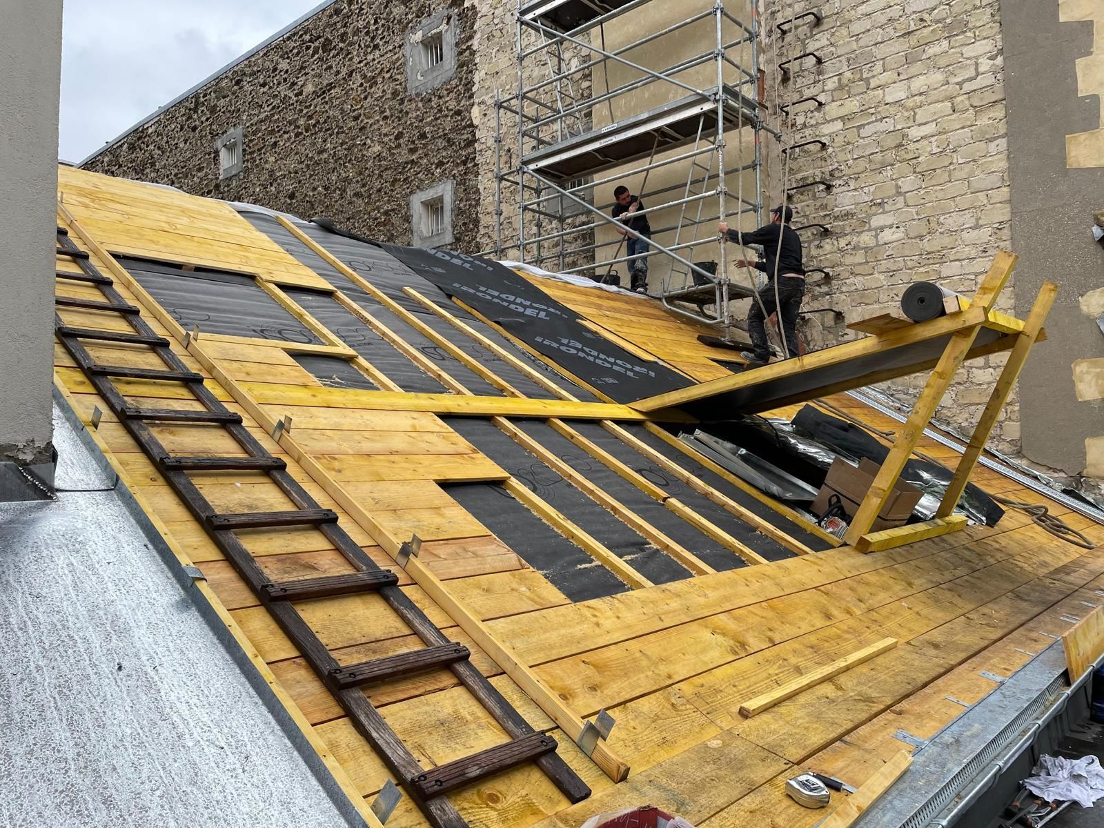 Des couvreurs posent une sous-couche noire sur un toit en pente.