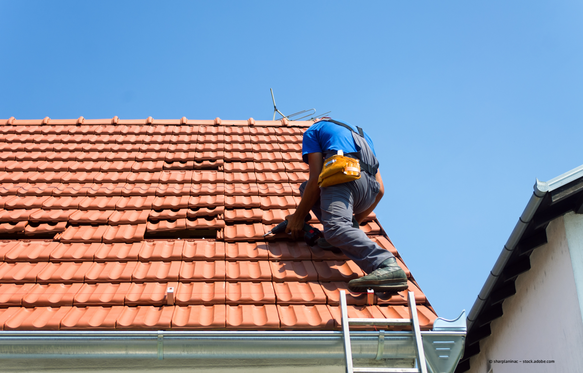 Ein Arbeiter repariert auf einer Leiter rote Dachziegel unter einem klaren blauen Himmel