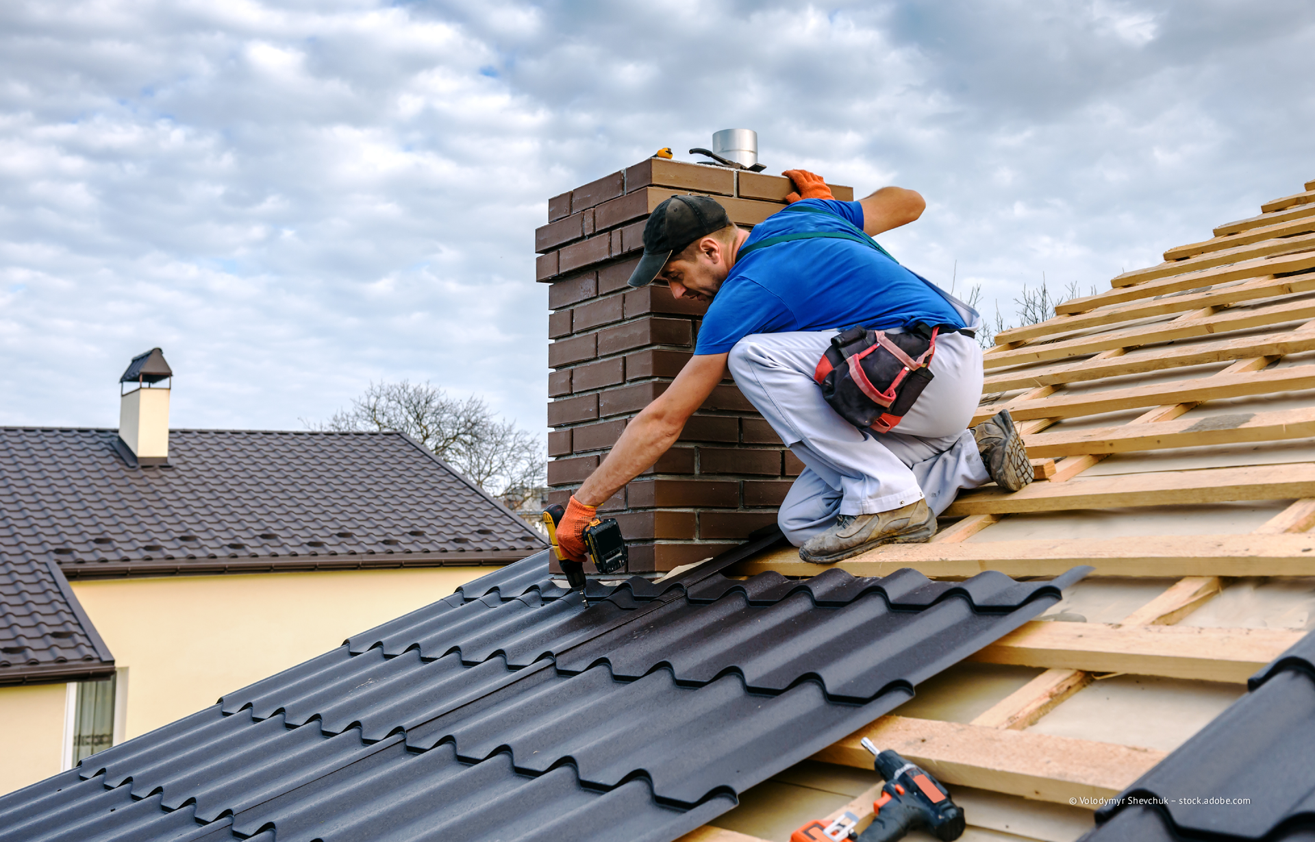 Ein Arbeiter verlegt Dachschindeln neben einem gemauerten Schornstein auf dem Dach eines Hauses.