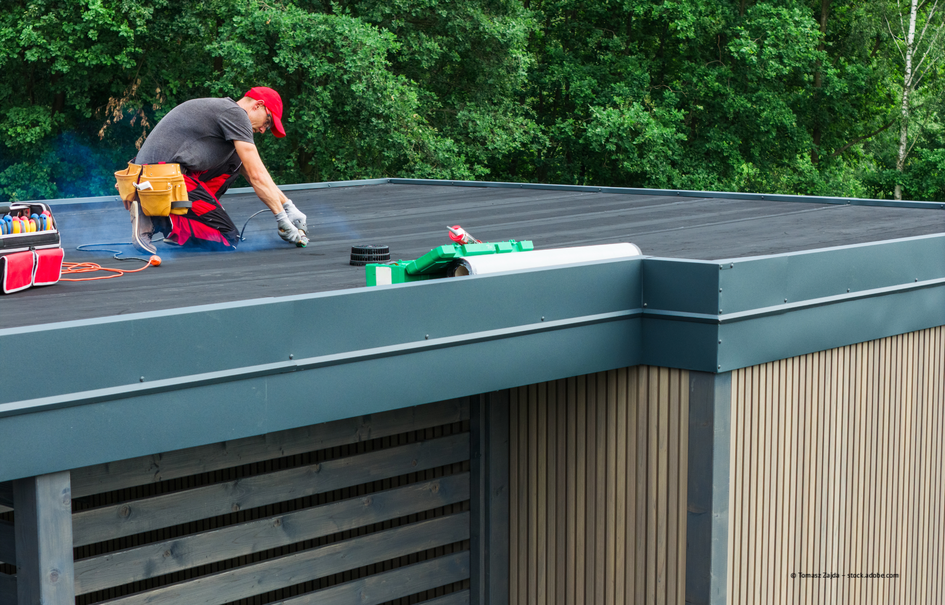 Ein Arbeiter, der auf dem Dach eines Flachdachgebäudes kniet und eine rote Kappe trägt, installiert dort die Dacheindeckung.