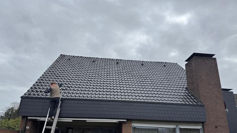 Ein Arbeiter repariert auf einer Leiter das graue Ziegeldach eines Hauses neben einem gemauerten Schornstein.