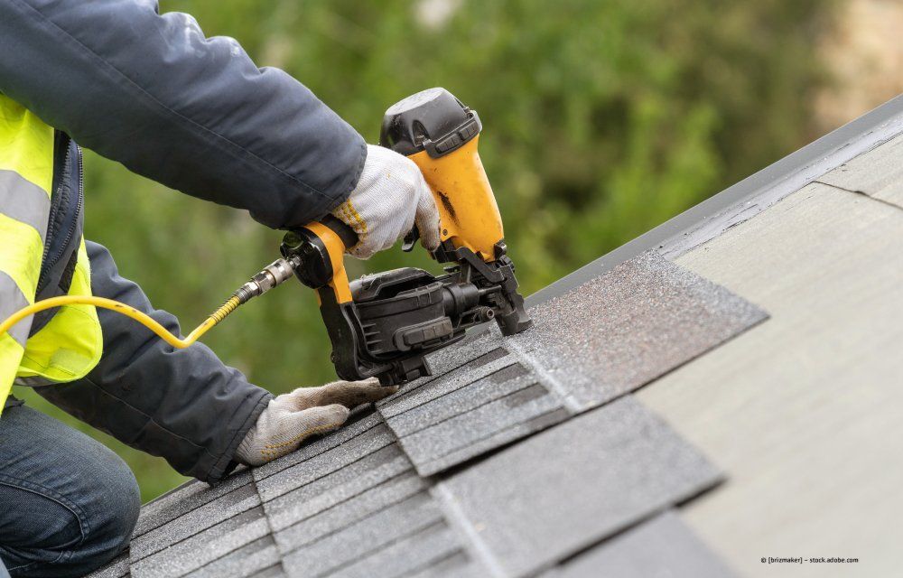 Ein Arbeiter befestigt mit einem Dachnagler graue Dachschindeln auf einem schrägen Dach.