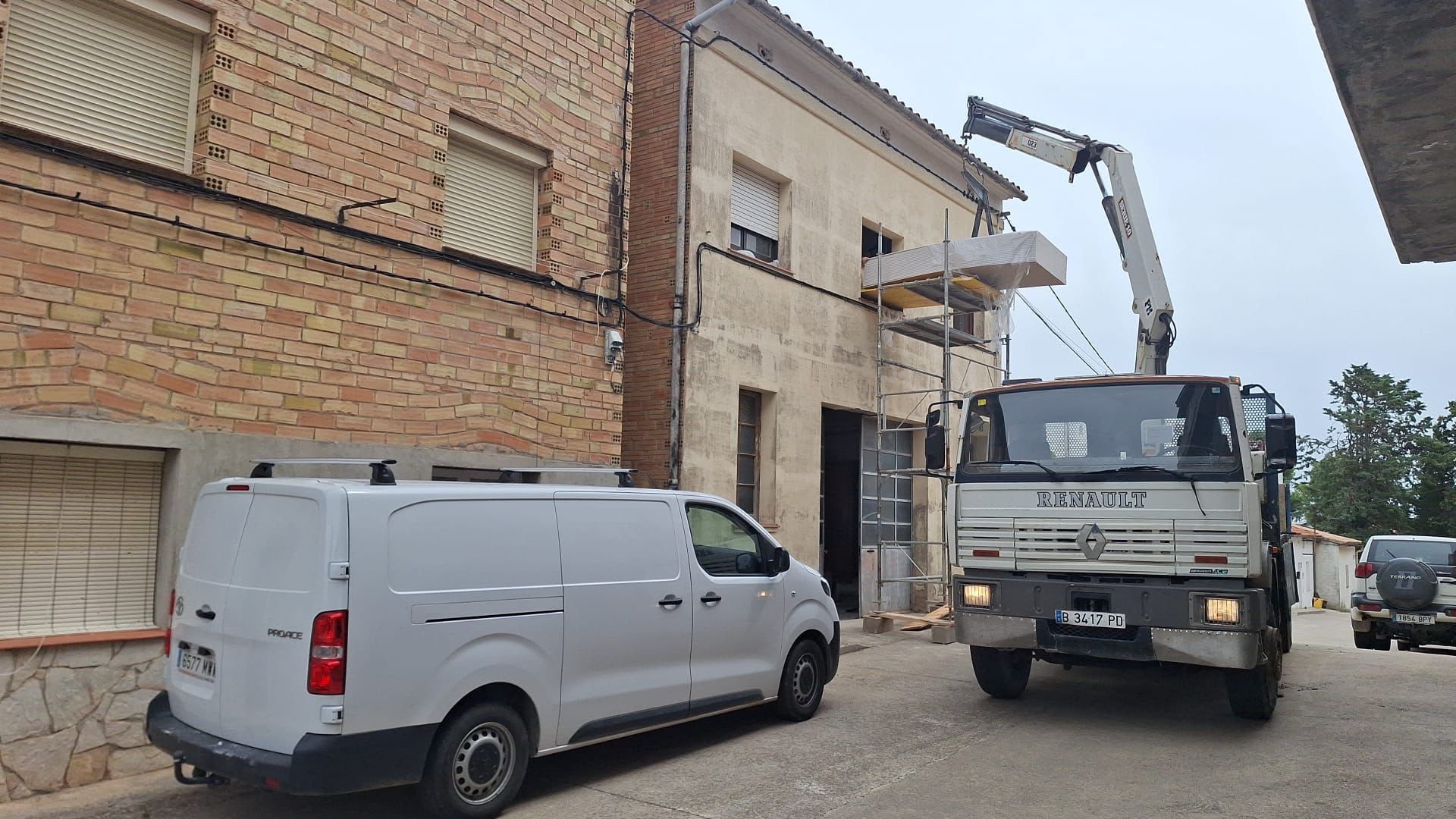 Una furgoneta blanca y un camión grúa estacionados junto a un edificio de ladrillos durante las obras de construcción.