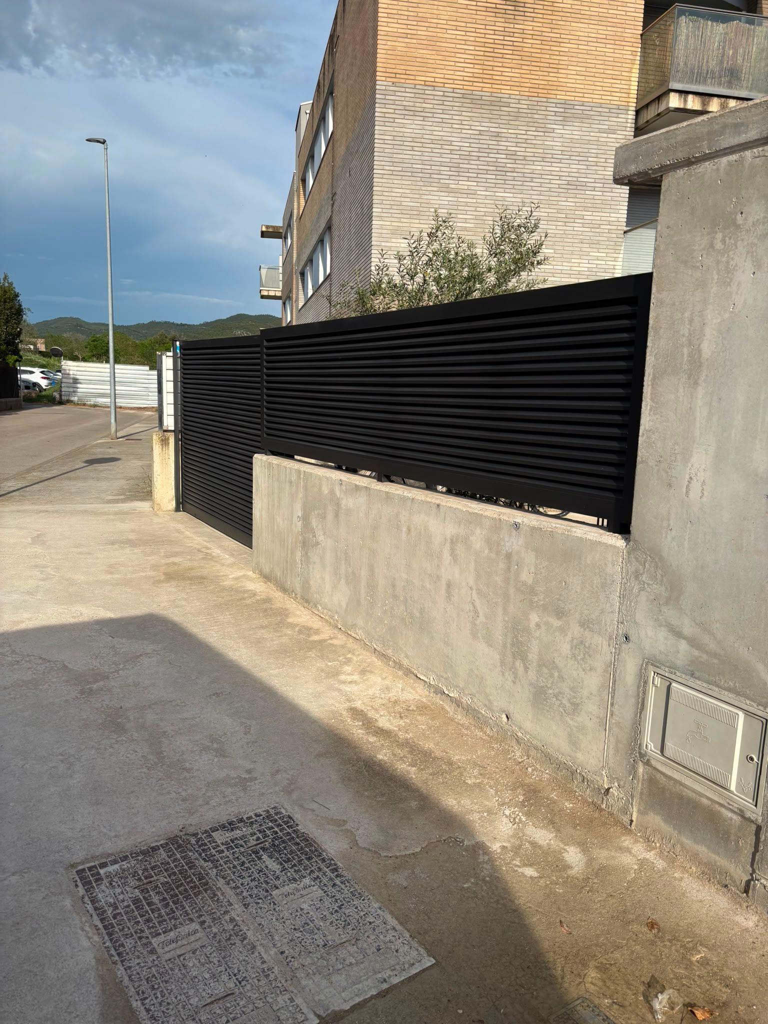 Calle residencial con una valla de listones negros junto a un muro de hormigón y una acera bajo un cielo nublado.