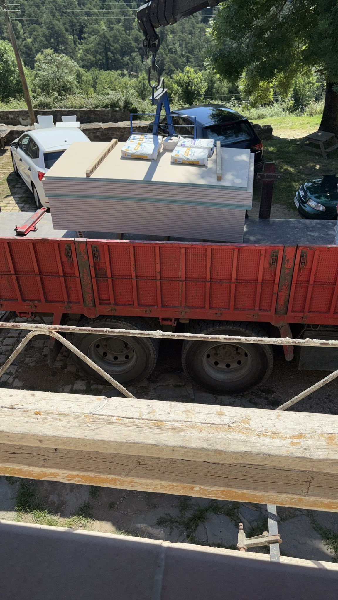 Camión volquete rojo cargado con paneles blancos apilados en una carretera junto a árboles y vigas de madera.