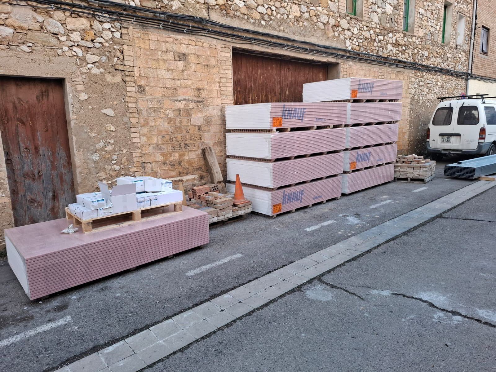 Montones de materiales envueltos en papel rosa en una calle junto a un muro de piedra, con una furgoneta