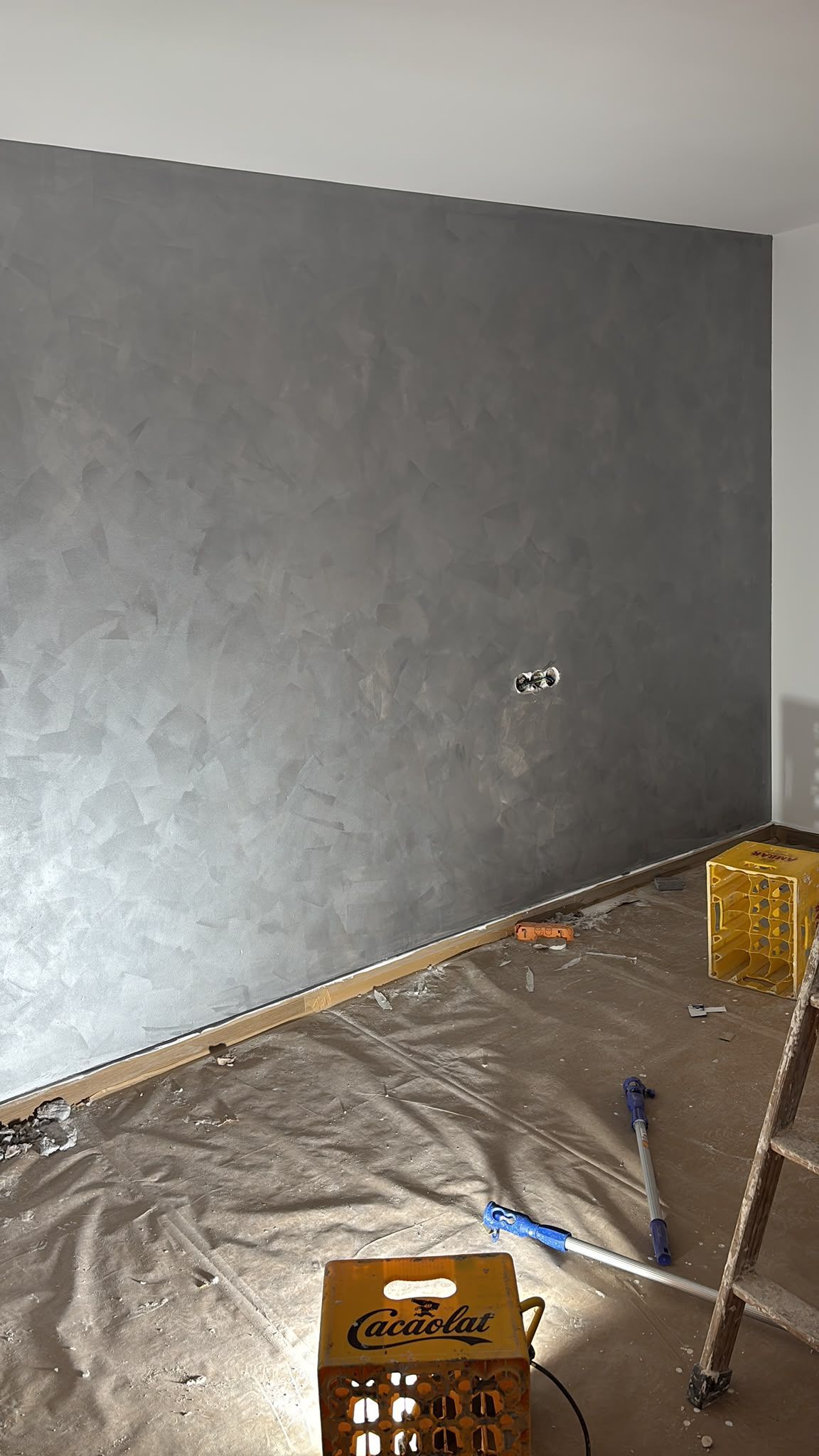 Pared gris en una habitación en obras, con suelo de plástico, herramientas y una escalera.